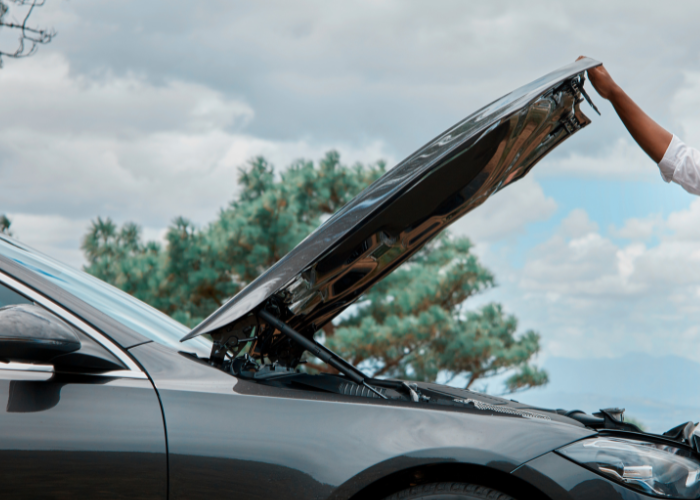 black car with lifted hood and person holding the hood with blue clouded sky and trees.
