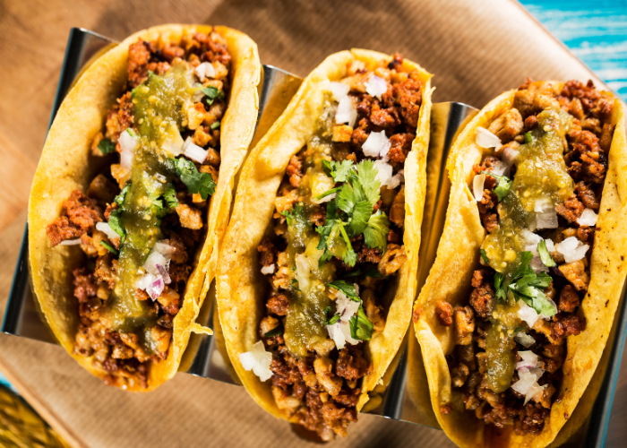 Hard shell tacos with meat cheese onions salsa in them on metal taco stand.