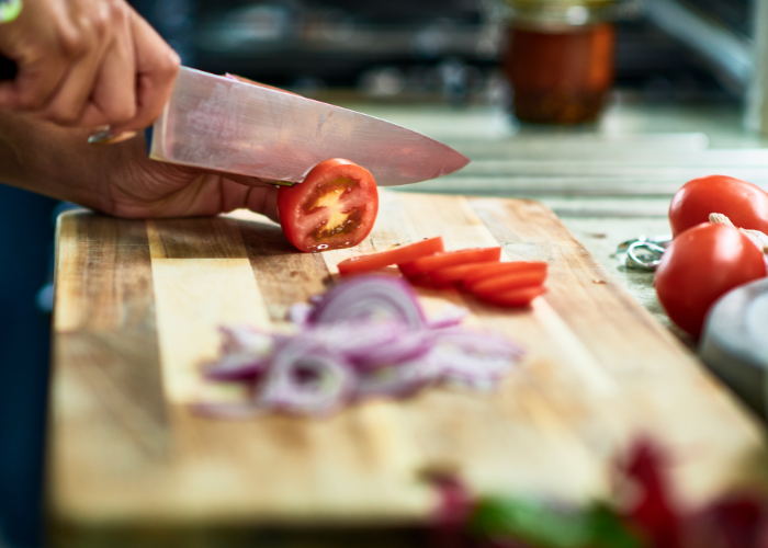 Woman slicing red tomatoes and red onion on light wood cutting board.