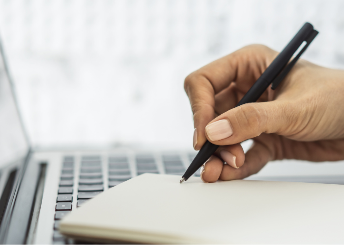 Woman reporter writing on notepad with pen in front of lap top.