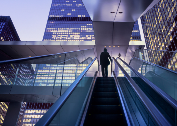 Man going up escalator during evening in front of lit up skyscrapers.