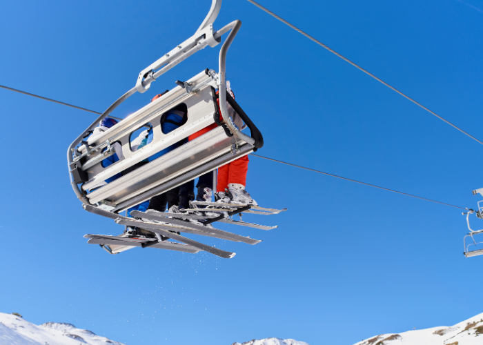 People skiing on mountain traveling by ski lift with blue sky and snowy mountains behind them.